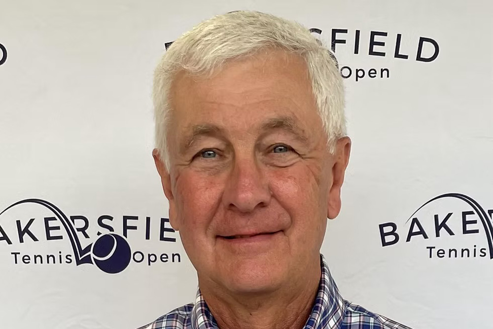 A headshot of Jerry Matthews smiling against a white step-and-repeat backdrop patterned with Bakersfield Tennis Open logos. He has short white hair and is wearing a blue, white, and red plaid button-down shirt.