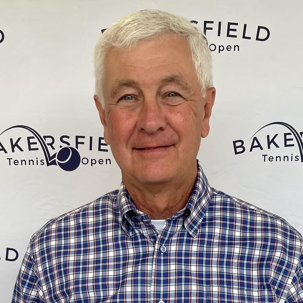 A headshot of Jerry Matthews smiling against a white step-and-repeat backdrop patterned with Bakersfield Tennis Open logos. He has short white hair and is wearing a blue, white, and red plaid button-down shirt.