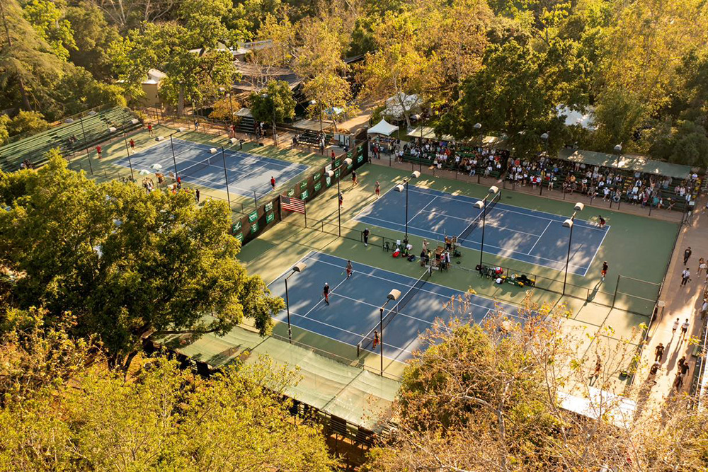 An aerial view of the tennis courts at Libbey Park bathed in warm afternoon sunlight during The Ojai Tennis Tournament. The blue hard courts with green surrounds are nestled beneath a dense canopy of large oak trees. Active matches are underway across multiple courts as spectators watch from shaded bleachers along the perimeter and walk along the park pathways.