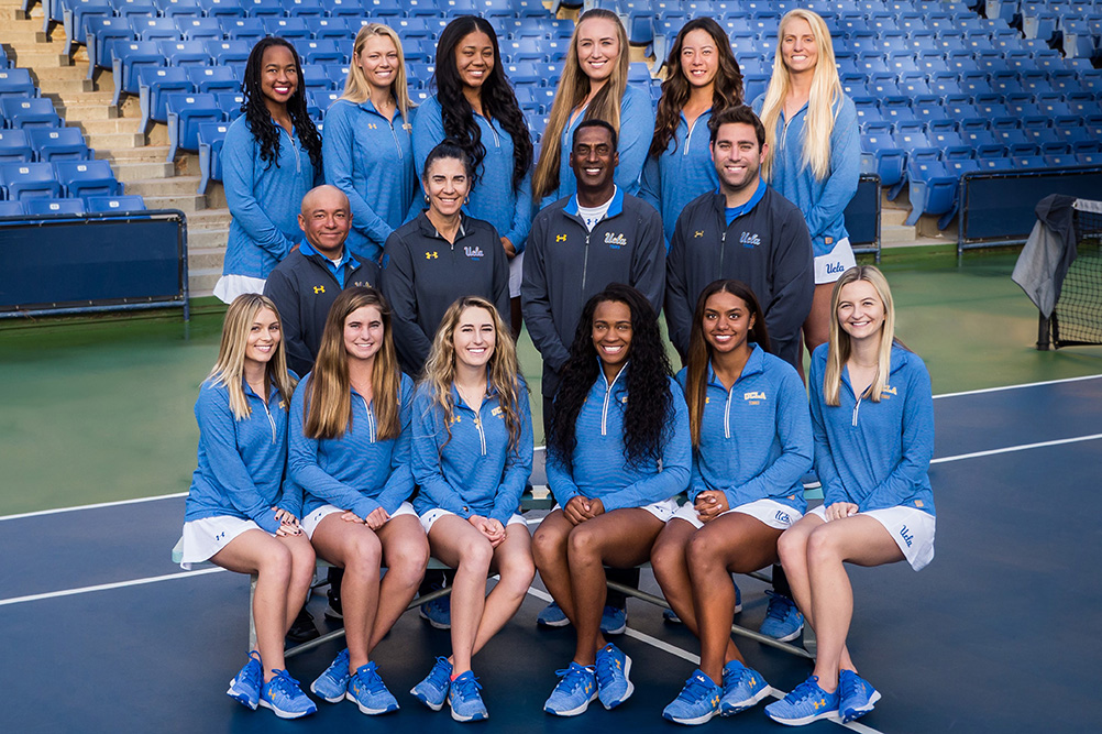 The 2018 UCLA Women's Tennis team and coaching staff pose in a long line across a hard court for Senior Day. The student-athletes wear matching grey long-sleeve shirts and white skirts, with senior players holding yellow flower bouquets in the center of the row in front of large stadium bleachers.