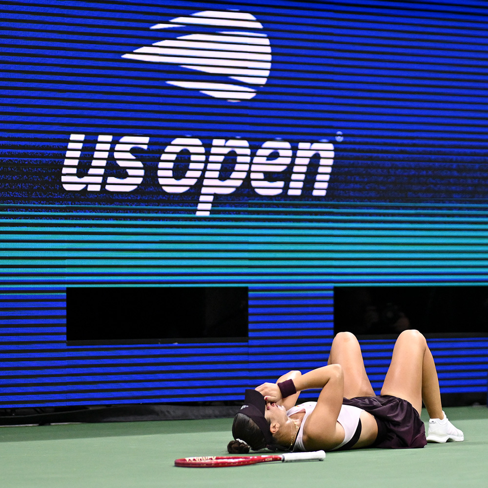 Tennis player Iva Jovic lies on her back on the court in emotional celebration after winning her third-round match at the US Open. She is resting flat on the green hard court with her knees bent, white shoes flat on the ground, and hands covering her face. Her red tennis racket lies on the court right beside her. Above her, a massive digital LED wall displays the white US Open logo against a vibrant blue-to-teal gradient background.