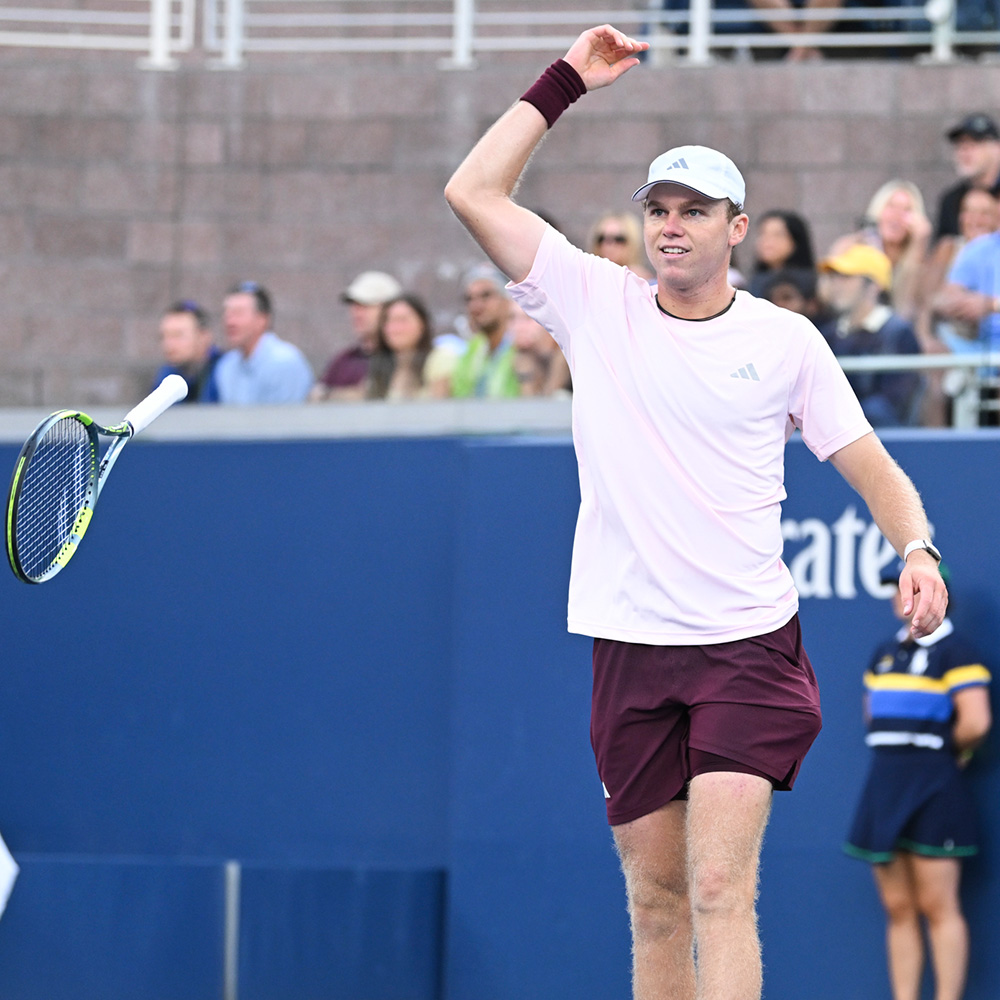 Alex Michelsen tossing his tennis racquet into the air as he celebrates winning match point at the US Open. Dressed in a light pink Adidas top, maroon shorts, white shoes, and a white cap, he raises his right arm in victory with blurred spectators watching from the stadium stands behind him.