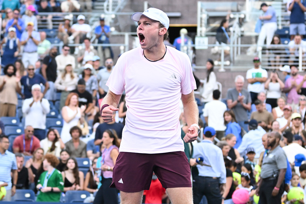 Alex Michelsen standing on the Grandstand blue hard court at the US Open, letting out an intense roar with double clenched fists in celebration after winning his fourth round match. He wears a light pink Adidas t-shirt, maroon shorts, white socks, white shoes, and a white baseball cap. In the background, his opponent walks toward the bench, ball persons move across the court, and thousands of spectators in the stadium stands cheer and applaud.