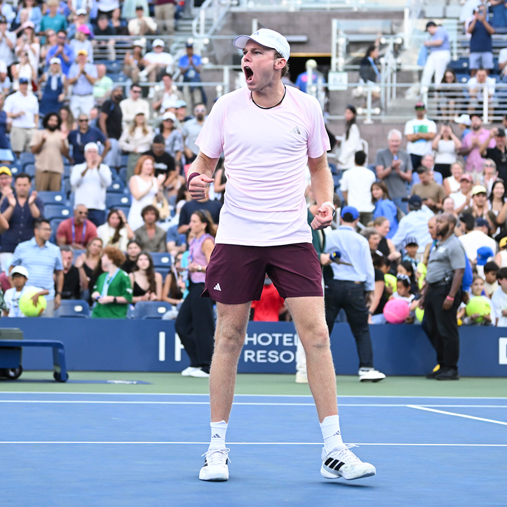 Alex Michelsen standing on the Grandstand blue hard court at the US Open, letting out an intense roar with double clenched fists in celebration after winning his fourth round match. He wears a light pink Adidas t-shirt, maroon shorts, white socks, white shoes, and a white baseball cap. In the background, his opponent walks toward the bench, ball persons move across the court, and thousands of spectators in the stadium stands cheer and applaud.