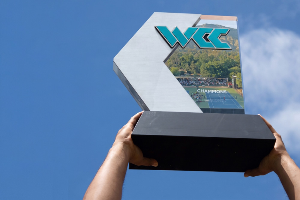 Two hands hold the West Coast Conference (WCC) championship trophy high overhead against a clear blue sky, celebrating the announcement that the WCC Tennis Championships will be coming to Libbey Park in 2027. The modern trophy features a dark rectangular base supporting a metallic silver and glass plaque topped with the turquoise and silver WCC logo, featuring an embedded image of tennis courts on the right.