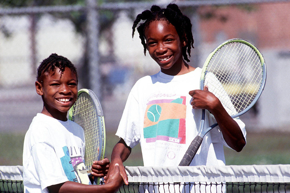 Young sisters Serena Williams (left) and Venus Williams (right) shake hands over a tennis net as children. Both smile brightly while holding their tennis rackets, wearing white graphic t-shirts on an outdoor public tennis court in Compton, California