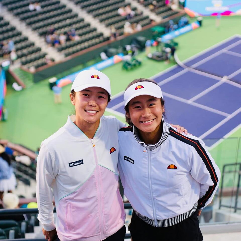 Former professional player turned coach Danielle Lao poses with young tennis star Natalie Kha in front of empty stadium seating overlooking an outdoor tennis court. Both wear white Ellesse caps and matching white jacket outerwear with the Ellesse logo.