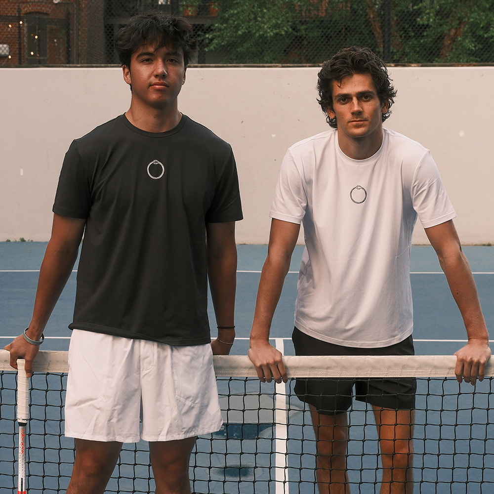 LET Tennis founders Jakob Esterowitz and Jonah Dickson stand side-by-side behind a tennis net on a blue court. Both wear t-shirts featuring the small, circular LET Tennis logo on the chest—one in a dark grey shirt and white shorts, and the other in a white shirt and black shorts. They look directly at the camera, with the grey court wall and background trees behind them.