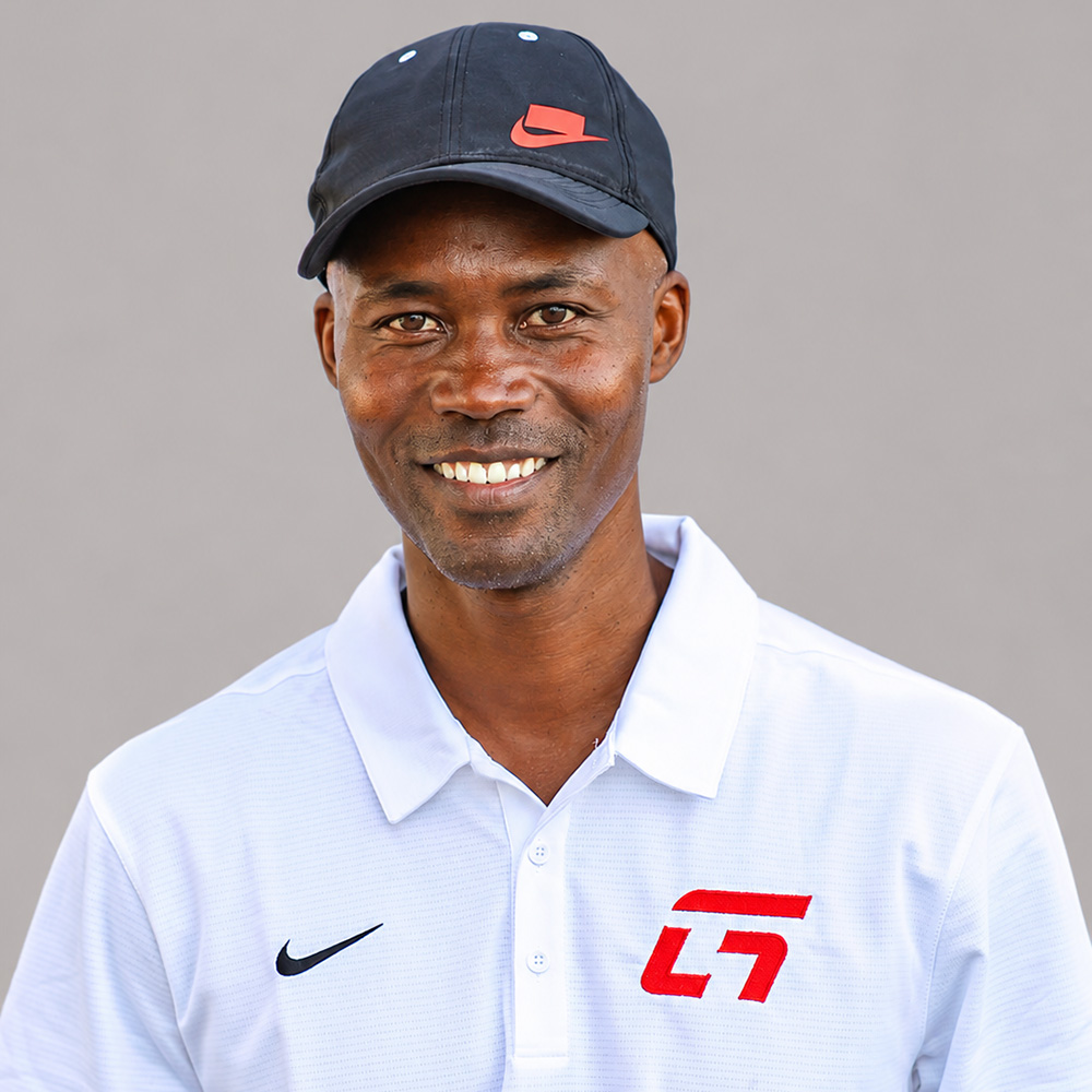 Headshot portrait of tennis coach Gerald Mathews, founder of Team G Tennis, smiling warmly directly at the camera against a neutral grey background. He wears a dark grey Nike baseball cap featuring a red swoosh box logo, along with a white Nike collared polo shirt adorned with a red "G" logo on the left chest.