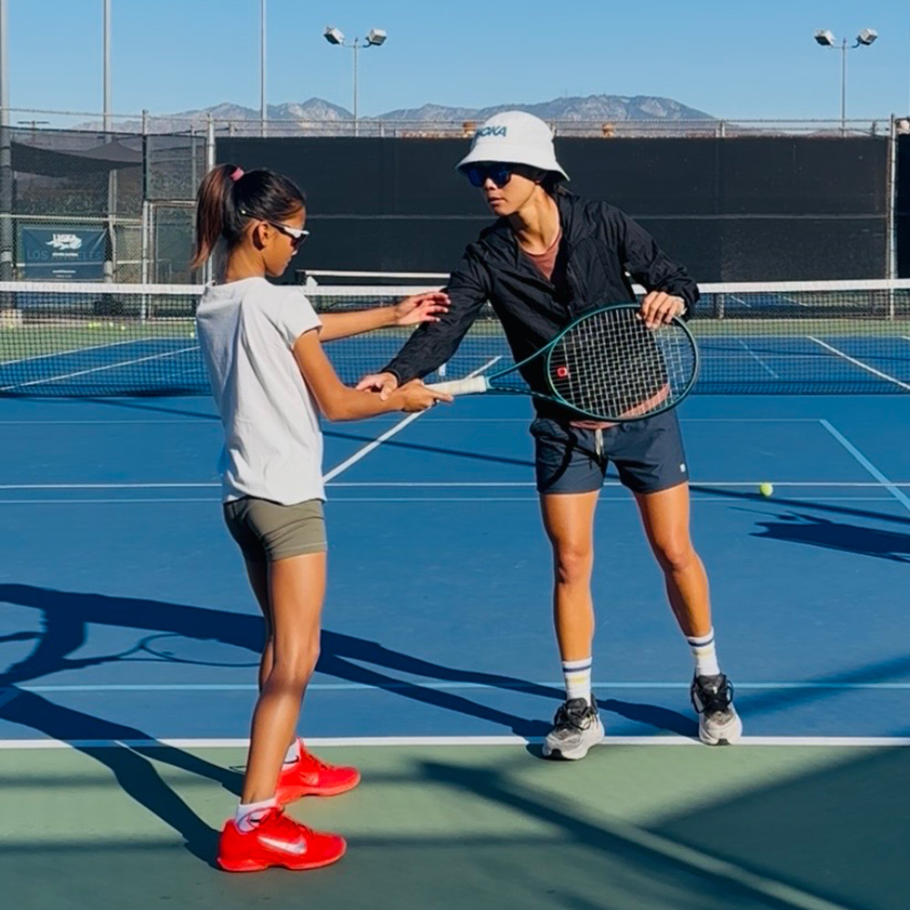 Tennis coach Danielle Lao gives hands-on instruction to a young player on a blue hard court under a clear sky. Lao wears a white Hoka bucket hat, sunglasses, a black jacket, dark shorts, and athletic shoes as she holds a tennis racket and guides the junior player's grip and arm positioning. The young player, dressed in a white shirt, green shorts, sunglasses, and bright red shoes, focuses on adjusting her stroke technique.