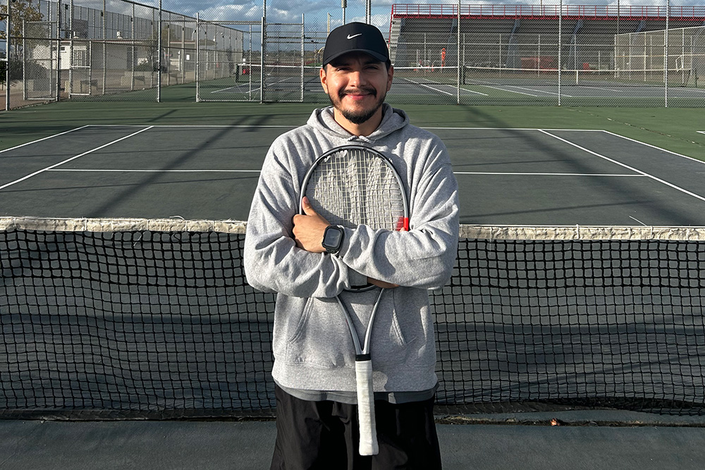 Tennis coach David Santos stands smiling on an outdoor tennis court with his arms crossed, holding a silver tennis racket vertically against his chest. He wears a black Nike baseball cap, a grey pullover hoodie, a black smartwatch, and black athletic shorts. He is positioned directly behind the court net, with empty green tennis courts and fenced court surrounds visible under a partly cloudy sky behind him.