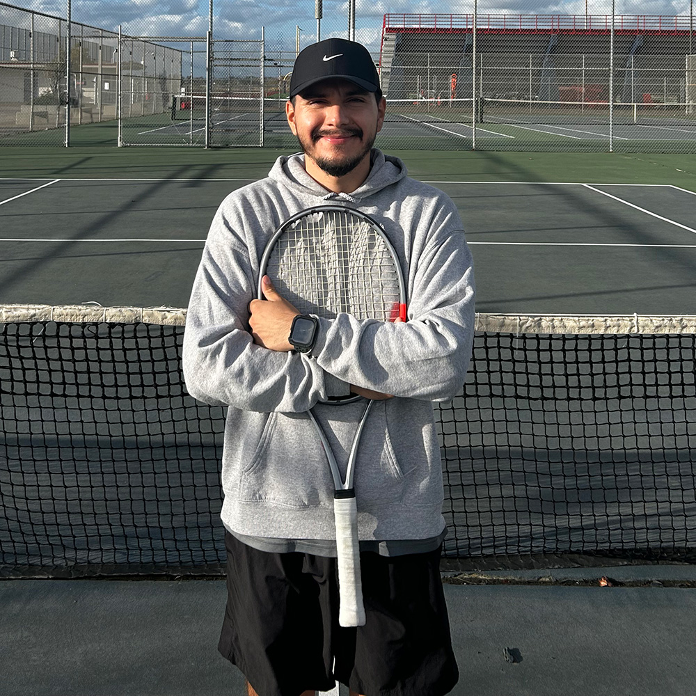 Tennis coach David Santos stands smiling on an outdoor tennis court with his arms crossed, holding a silver tennis racket vertically against his chest. He wears a black Nike baseball cap, a grey pullover hoodie, a black smartwatch, and black athletic shorts. He is positioned directly behind the court net, with empty green tennis courts and fenced court surrounds visible under a partly cloudy sky behind him.