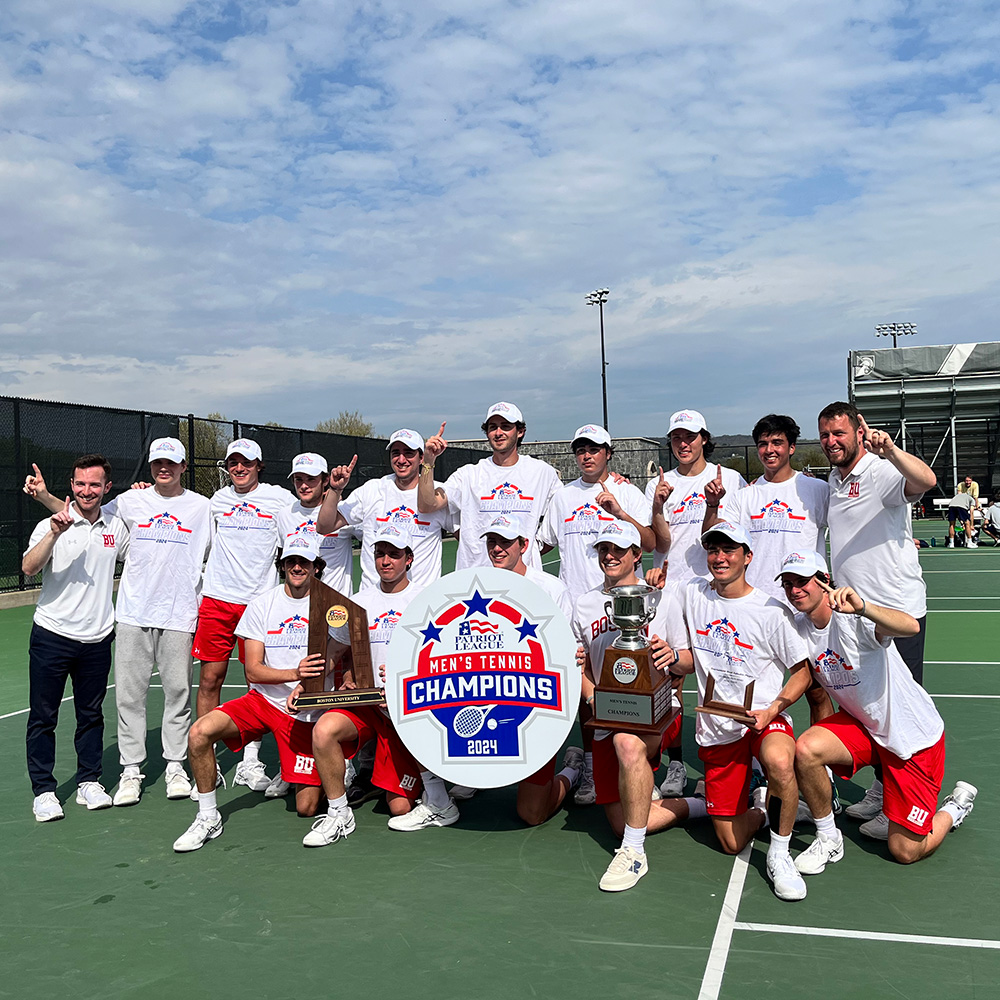 The Boston University Men's Tennis team, including LET Tennis founders Jonah Dickson and Jakob Esterowitz, celebrates on court after winning the 2024 Patriot League Championship Conference Title. The players and coaches pose together wearing matching white championship shirts, white hats, and red shorts. They hold up several tournament trophies and a large circular sign reading "Patriot League Men's Tennis Champions 2024," with many raising a finger to celebrate their number-one finish under a partly cloudy sky.