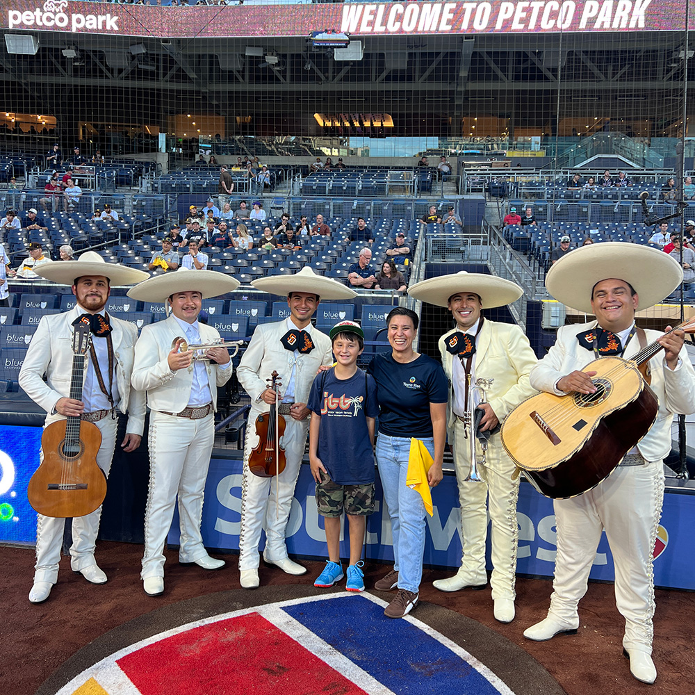 Gaby Millán and Michael Monzón stand on the warning track at Petco Park surrounded by five members of a traditional mariachi band wearing white charro suits and sombreros. The group poses smiling in front of the field boxes beneath a "WELCOME TO PETCO PARK" stadium sign.