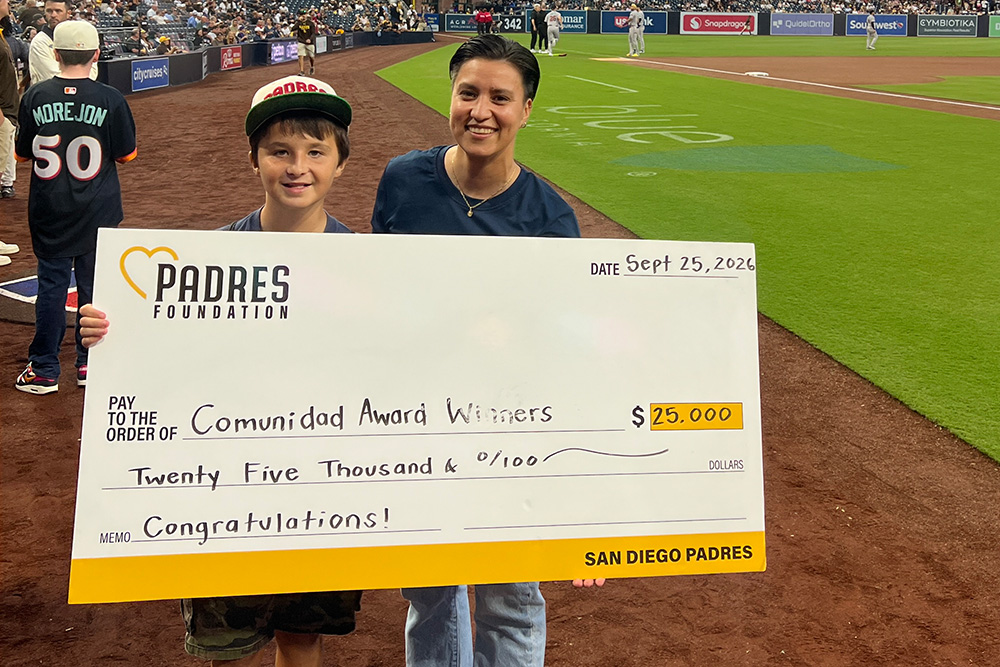 Teens Rise Foundation Executive Director Gaby Millán and youth participant Michael Monzón smile while standing on the field at Petco Park holding a ceremonial oversized check. The check, presented by the Padres Foundation, honors TRF and its South Bay Serves tennis program with the Comunidad Award for their impactful community work.