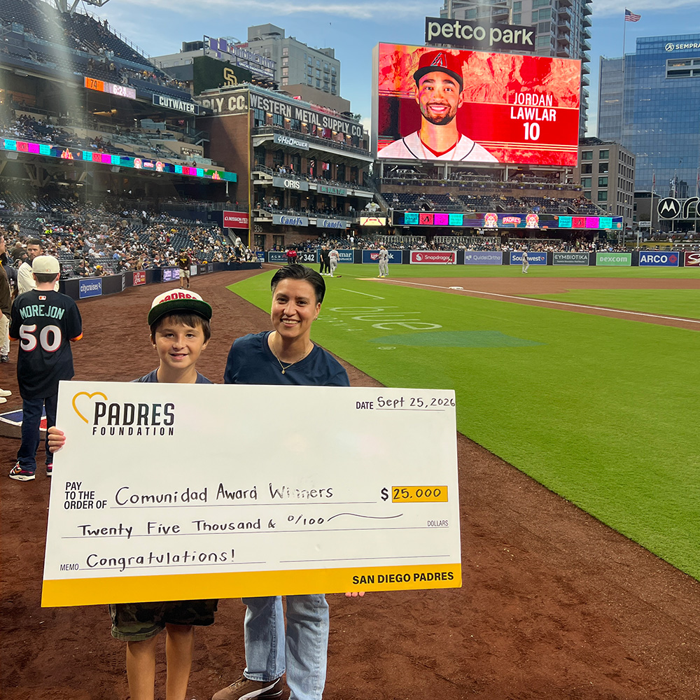 Teens Rise Foundation Executive Director Gaby Millán and youth participant Michael Monzón smile while standing on the field at Petco Park holding a ceremonial oversized check. The check, presented by the Padres Foundation, honors TRF and its South Bay Serves tennis program with the Comunidad Award for their impactful community work.