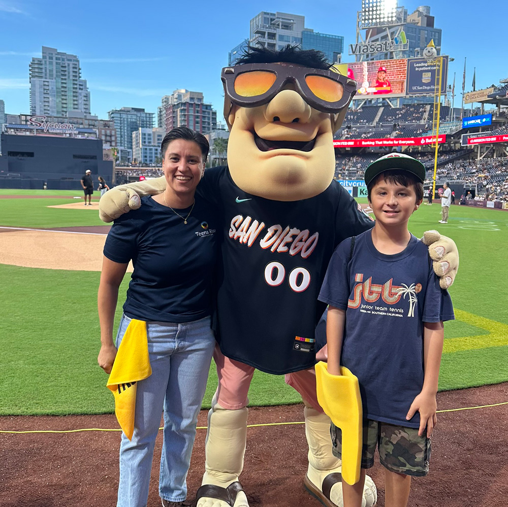 Gaby Millán and Michael Monzón pose on the outfield edge at Petco Park with the San Diego Padres mascot, the Swinging Friar, who puts his arms around both of them. Millan wears a navy Teens Rise Foundation shirt, and Mozon wears a navy USTA Junior Team Tennis t-shirt while holding yellow stadium rally towels.