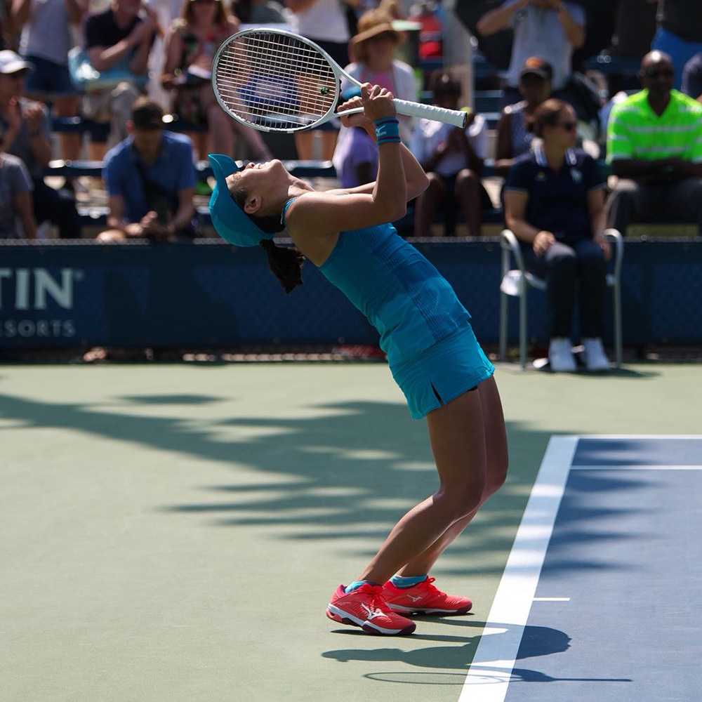 Professional tennis player Danielle Lao arching her back in an emotional celebration on the hard court after a point victory at the US Open. Dressed in an all-blue Mizuno outfit with matching cap and red tennis shoes, she holds her racket over her face while standing near the baseline.