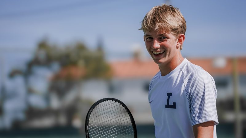 Braun Levi smiles while wearing a Loyola High School shirt and carrying a tennis racquet