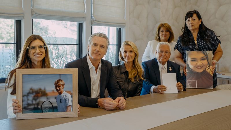 Jennifer Levi, California Governor Gavin Newsom, First Partner Jennifer Siebel Newsom, and victim advocates pose together at a conference table for the signing of "Braun's Law" (SB 907) and related DUI legislation. On the left, Levi holds a framed photograph of her late son, Braun Levi, wearing a tennis shirt on court. On the right, family members hold a portrait of another victim as the group sits together around a long wooden table.