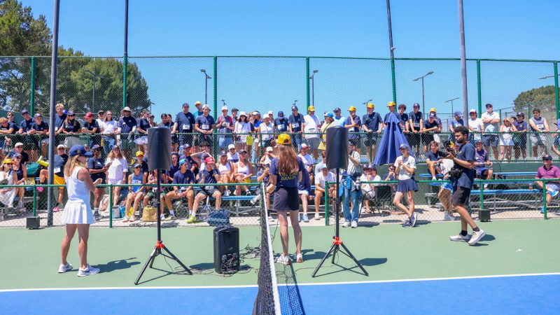 A wide view of the opening ceremony at the second annual Live Like Braun (LLB) Memorial Tennis Tournament. Two speakers address a large crowd of participants and supporters gathered along the court fence and bleachers. A PA system with speakers on tripods is set up near the net, while players in athletic wear listen under a bright, sunny sky on an outdoor blue and green hard court.